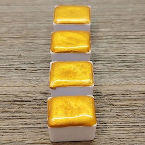 Op de afbeelding: Vier gouden aquarelverfblokken in witte plastic bakjes zijn in een verticale rij gerangschikt. De metallic gouden verf heeft een glanzend, reflecterend oppervlak. De bakjes staan tegen een getextureerde, lichtbruine houten achtergrond.