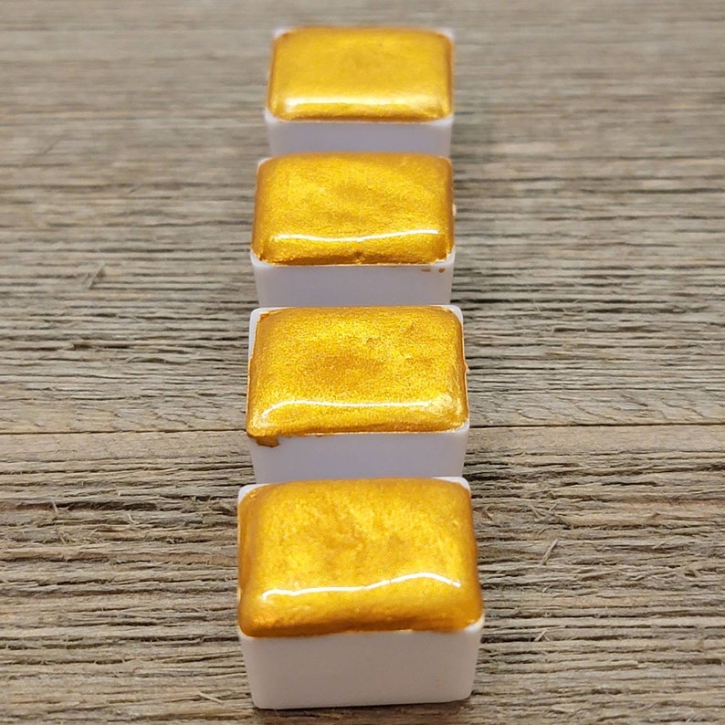 Op de afbeelding: Vier gouden aquarelverfblokken in witte plastic bakjes zijn in een verticale rij gerangschikt. De metallic gouden verf heeft een glanzend, reflecterend oppervlak. De bakjes staan tegen een getextureerde, lichtbruine houten achtergrond.