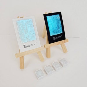 May include: Two small wooden easels display watercolor swatches. One is on white paper, the other on black, both with the word "Unicorn." Below are four small, rectangular watercolor pans. The swatches and pans have a shimmering, iridescent quality.