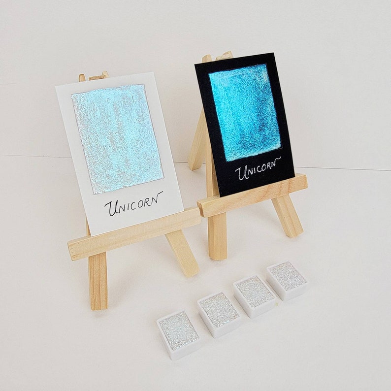May include: Two small wooden easels display watercolor swatches. One is on white paper, the other on black, both with the word "Unicorn." Below are four small, rectangular watercolor pans. The swatches and pans have a shimmering, iridescent quality.