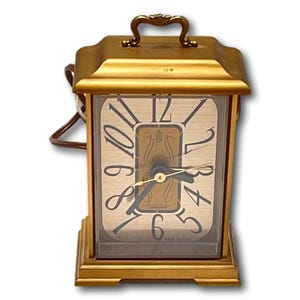 May include: A vintage gold-toned electric clock with a rectangular shape. The clock face has black numerals and hands, with a decorative center design. It features a handle on top and a power cord.