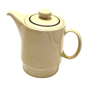 May include: A cream-colored ceramic teapot with a speckled finish, featuring a ribbed base, a curved handle, and a matching lid with a dark brown circular accent line and a small, ridged knob handle on top for easy removal and secure placement.
