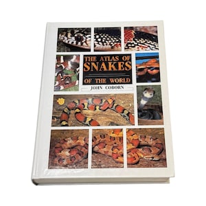 May include: A hardback book titled The Atlas of Snakes of the World by John Coborn. The white cover features a collage of various snake species in red, black, and orange patterns, including a cobra and a coiled snake on a textured ground.