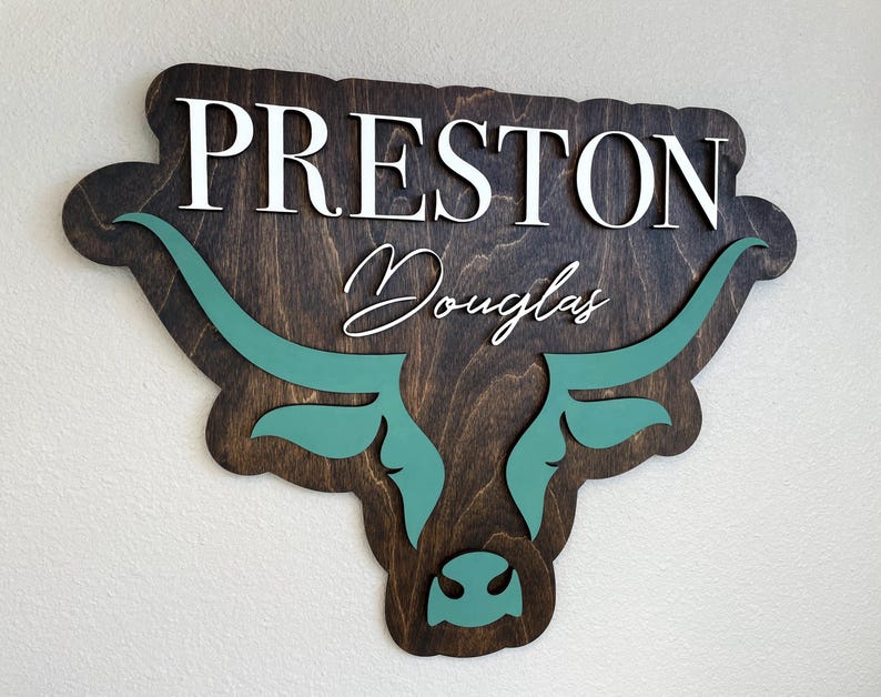 May include: Wooden sign featuring the name "PRESTON" in white capital letters above the name "Douglas" in cursive. The sign is shaped like a bull's head, with teal horns and facial details against a dark brown background.
