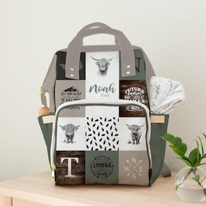 May include: A multi-colored diaper bag with a patchwork design. The bag features images of Highland cows, text that reads "Noah Tate", and other decorative elements. The bag has multiple pockets and a handle.