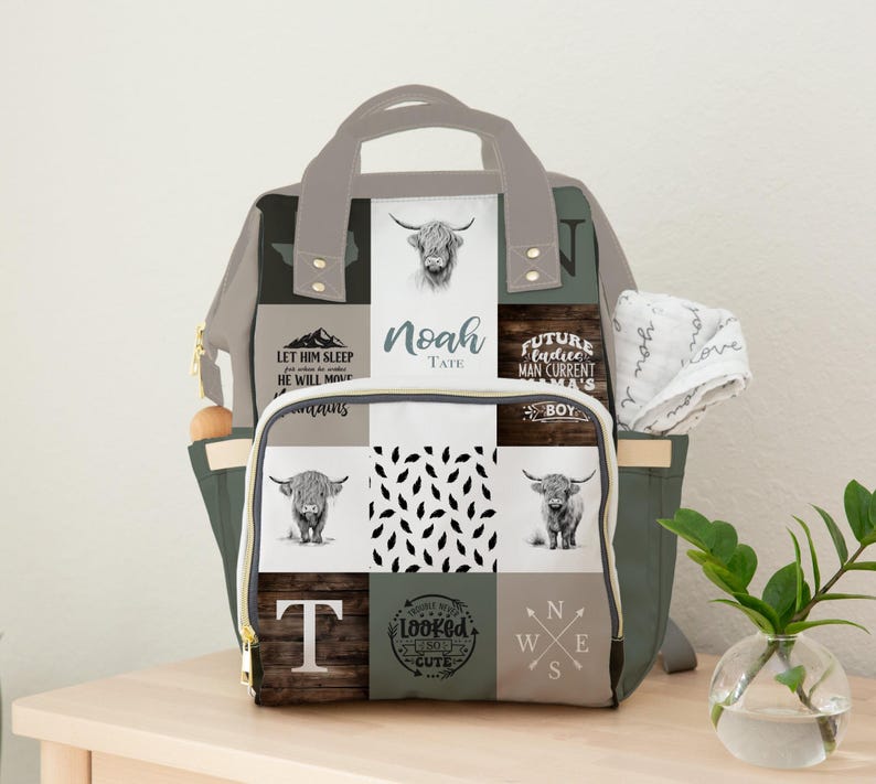 May include: A multi-colored diaper bag with a patchwork design. The bag features images of Highland cows, text that reads "Noah Tate", and other decorative elements. The bag has multiple pockets and a handle.