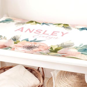 May include: White changing pad cover with a floral design featuring pink and green watercolor flowers and leaves. The text "ANSLEY Evelyn" is printed in pink on the cover.