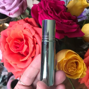May include: A shiny, silver perfume bottle is held against a backdrop of vibrant roses in various colours. The roses include shades of pink, red, orange, and yellow. The bottle has a cylindrical shape and a cap.