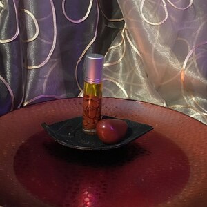 Puede incluir: Una pequeña botella de vidrio de aceite esencial con tapa plateada, llena de líquido amarillo y elementos decorativos marrones, se encuentra sobre un plato negro en forma de hoja. Una piedra roja en forma de corazón descansa al lado.