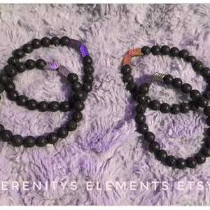 Può includere: Due braccialetti con perline in pietra lavica nera con accenti chevron metallici. Uno ha uno chevron argentato, l'altro ha uno chevron rame e argento. I braccialetti sono esposti su una superficie morbida e strutturata. Il testo "SERENITYS ELEMENTS ETSY" è in basso.