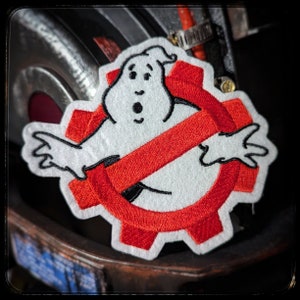 May include: Red and white embroidered patch with a ghost and a red slash through it. The ghost is white with black eyes and a mouth. The patch is shaped like a gear.