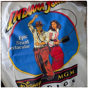 May include: White t-shirt featuring a graphic of Indiana Jones and a woman, with the text "INDIANA JONES" in yellow and red. The graphic is set against a blue background with the words "Epic Stunt Spectacular".
