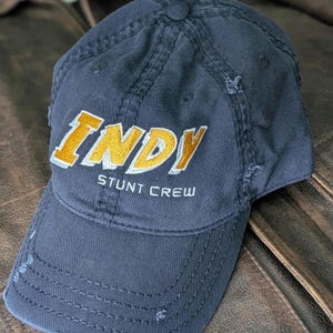 May include: A navy blue baseball cap featuring the word "INDY" in gold and white embroidery, with "STUNT CREW" underneath. The cap has a distressed appearance with worn edges.