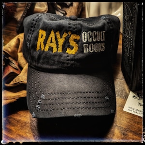 Può includere: Cappellino da baseball nero con dettagli usurati. Il cappellino presenta la scritta "RAY'S" in giallo e "OCCULT BOOKS" in bianco. Il cappello è realizzato in un tessuto resistente e ha una visiera curva.