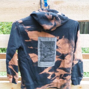 May include: A black hooded sweatshirt featuring a bleached, mottled pattern of orange-brown patches throughout. A rectangular grey fabric patch is sewn onto the center back, displaying a black-and-white graphic illustration of a moth.