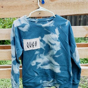 May include: A long-sleeve crewneck sweatshirt in a teal blue color with a bleached tie-dye pattern. A rectangular off-white fabric patch is sewn onto the upper left chest, featuring the word OUCH printed in a stylized black gothic font.