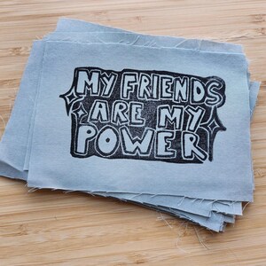 May include: A stack of light blue fabric squares with the text "MY FRIENDS ARE MY POWER" in black block letters. The text is inside a decorative frame. The squares are on a wooden surface.