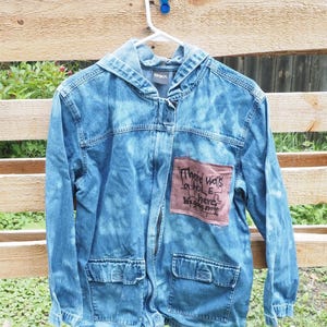 May include: A blue denim hooded jacket with a bleached wash effect, featuring a brown rectangular patch on the chest with the handwritten text: There was a hole here, it's gone now. The jacket includes two front flap pockets and a front zipper closure.