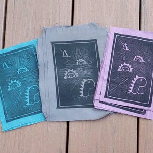 May include: Three rectangular fabric patches featuring a block print design of a dinosaur and suns. The patches are teal, gray, and light purple, each with a dark ink print enclosed in a rectangular border, showcasing a stylized prehistoric creature.