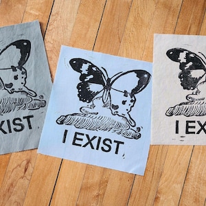 May include: Three fabric squares with a black and white butterfly graphic and the text "I EXIST" printed in black. The squares are in different shades of blue, gray, and white.