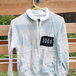 May include: A light blue and white tie-dye quarter-zip sweatshirt with a high collar and long sleeves. A rectangular black patch is sewn onto the left chest featuring the white text OUCH in a stylized, jagged font. The garment has two front pockets.