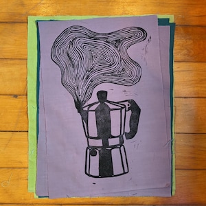 May include: A rectangular piece of light purple fabric featuring a black ink block print of a moka pot with stylized, swirling steam rising from the top. The fabric is layered over additional pieces of green and dark teal fabric.