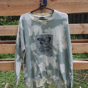 May include: A long-sleeve crewneck sweatshirt featuring a sage green and white tie-dye pattern. A rectangular patch is centered on the chest, displaying a black graphic of a butterfly above the text I EXIST in a bold, sans-serif font.