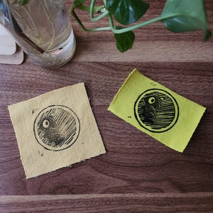 May include: Two yellow fabric patches with black ink printed designs. The designs are circular and feature a stylized image of a creature with a large eye.