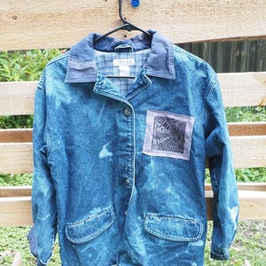May include: A blue denim jacket with a bleached, mottled wash, featuring a dark blue collar and a plaid interior lining. A square patch on the chest displays a spiderweb graphic with the text "GOOD NIGHT" printed in black ink on a light fabric.