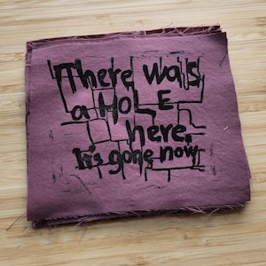 May include: A square, mauve fabric with black text that reads "There was a hole here. It's gone now." The text is in a blocky, artistic font. The fabric has a raw, unfinished edge.