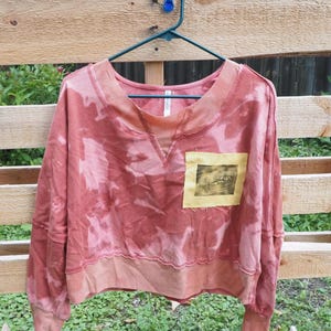 May include: A long-sleeve pullover sweatshirt in a faded coral and pink tie-dye pattern. The garment features a wide ribbed neckline with a V-stitch detail, a wide ribbed hem, and a square yellow patch on the chest with a dark graphic print.