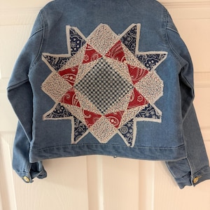 May include: A light blue denim jacket with a patchwork star design on the back. The star is made of red, white, and blue patterned fabric. The jacket has a collared neckline and gold buttons.