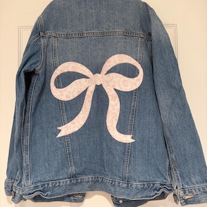 May include: A blue denim jacket with a large pink bow design on the back. The bow is made of a light pink fabric with a floral pattern. The jacket has a classic collar, button closures, and pockets.