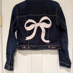 May include: A dark indigo denim jacket featuring a large, pale pink bow motif on the back. The jacket has a button-up front, two chest pockets, and buttoned cuffs. The bow is a key design element, adding a touch of femininity to the casual denim style.