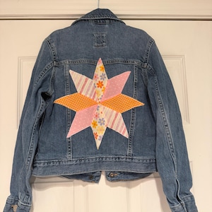 May include: A denim jacket with a large, colorful star design on the back. The star is made of various patterned fabrics, including pink polka dots, orange polka dots, and floral prints. The jacket is hanging on a metal hanger.