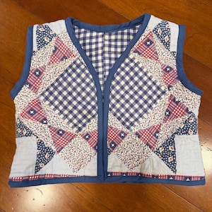 May include: A vintage quilted vest featuring a patchwork design with red, white, and blue fabrics. The vest has a blue gingham interior and is trimmed with blue binding. The overall style is reminiscent of Americana.