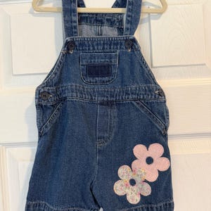 May include: Blue denim overall shorts with adjustable straps and button closures. Features two pink flower appliqués on the front. The garment is hanging on a wooden hanger.