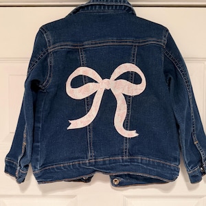 May include: A dark blue denim jacket with a large, light pink bow applique on the back. The bow has a delicate floral pattern. The jacket has a classic collar, button closures, and pockets.