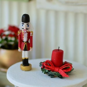 Könnte beinhalten: Eine Miniatur-Weihnachtsszene mit einem rot-goldenen Nussknacker und einer roten Kerze in einem Kranz. Der Nussknacker hat einen schwarzen Hut und weißes Haar. Die Kerze ist von grünen Blättern und einer roten Schleife umgeben.