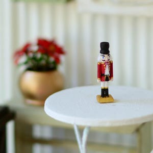 Puede incluir: Una figura de cascanueces de madera en miniatura. La figura viste una chaqueta roja, pantalones negros y un sombrero alto negro. Se encuentra sobre una pequeña base cuadrada de color dorado. El cascanueces está colocado sobre una mesa redonda blanca.