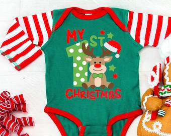 May include: Green and red striped long-sleeve baby onesie with the text "My 1st Christmas" and a cartoon reindeer with a red Santa hat. A gingerbread man toy with a candy cane is on the right side of the onesie.