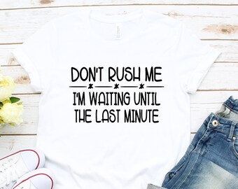 May include: White t-shirt with black text that reads "Don't rush me - I'm waiting until the last minute". The shirt is laid out on a white wooden surface with a pair of white sneakers and blue jeans.