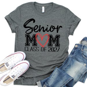 Op de afbeelding: Een donkergrijs gem&ecirc;leerd T-shirt met korte mouwen en de zwarte tekst Senior MOM, waarbij een rood hart de letter O vervangt, en de tekst CLASS OF 2027 eronder. Het T-shirt is gecombineerd met witte canvas sneakers en een blauwe spijkerbroek.