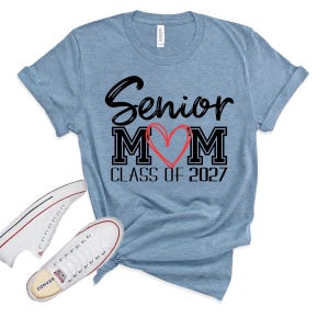 Op de afbeelding: Een gem&ecirc;leerd lichtblauw t-shirt met korte mouwen met de zwarte tekst Senior MOM, waarbij een handgetekend rood hart de letter O vervangt, en de tekst CLASS OF 2027 eronder. Een paar witte lage canvas sneakers liggen ernaast.