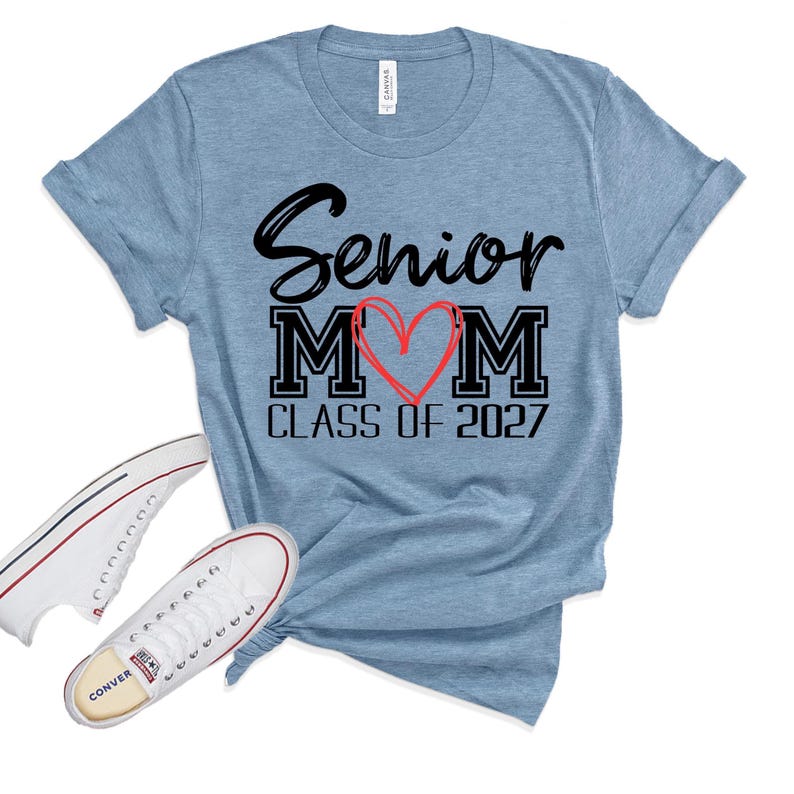 Op de afbeelding: Een gem&ecirc;leerd lichtblauw t-shirt met korte mouwen met de zwarte tekst Senior MOM, waarbij een handgetekend rood hart de letter O vervangt, en de tekst CLASS OF 2027 eronder. Een paar witte lage canvas sneakers liggen ernaast.