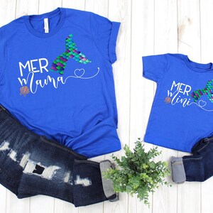 May include: Two royal blue t-shirts with a mermaid tail graphic and the text "Mer mama" and "Mer mini" on them. The shirts are laid out on a white wooden surface with a pair of blue jeans and a potted plant.