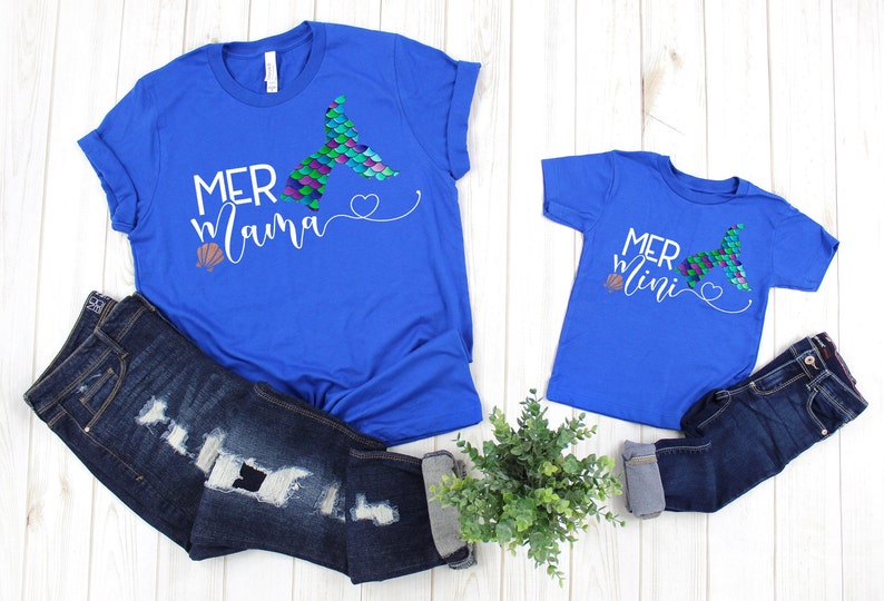 May include: Two royal blue t-shirts with a mermaid tail graphic and the text "Mer mama" and "Mer mini" on them. The shirts are laid out on a white wooden surface with a pair of blue jeans and a potted plant.