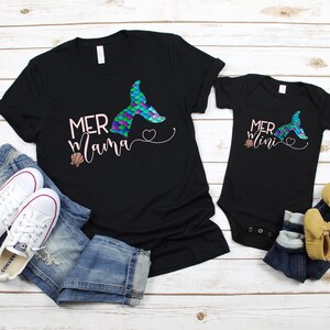 May include: A black t-shirt with the text "Mermama" and a mermaid tail graphic. A black bodysuit with the text "Mermini" and a mermaid tail graphic. Both shirts have a pink heart and a seashell graphic.
