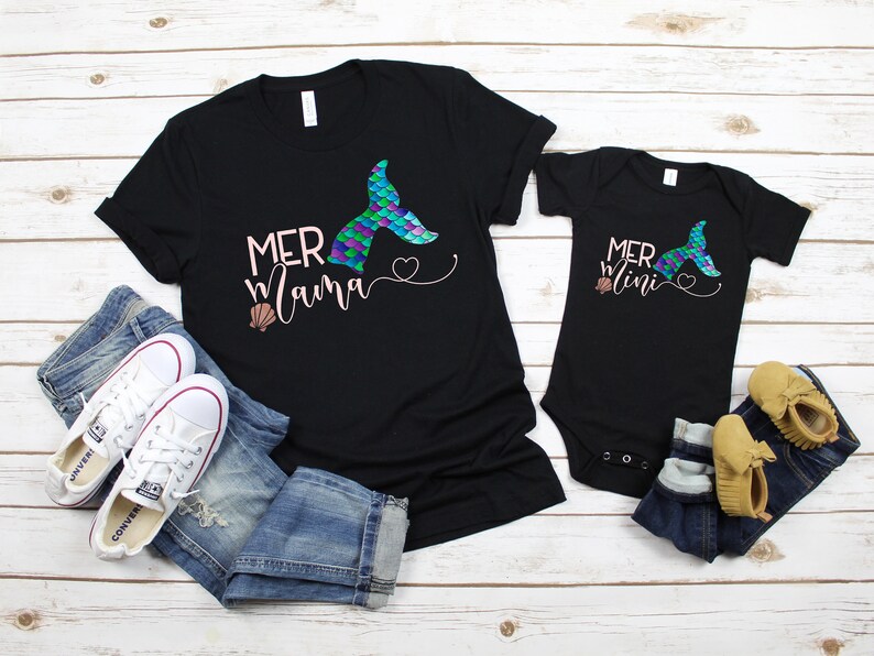 May include: A black t-shirt with the text "Mermama" and a mermaid tail graphic. A black bodysuit with the text "Mermini" and a mermaid tail graphic. Both shirts have a pink heart and a seashell graphic.