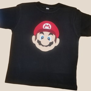 Op de afbeelding: Zwart T-shirt met een rode en witte Mario-personage afbeelding.